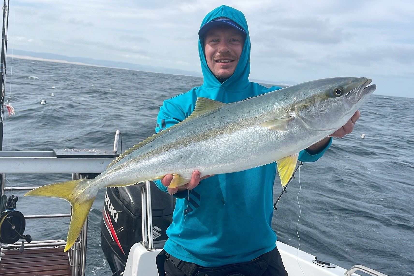Smiling angler holding a yellowtail across his chest, outboard and coastline behind